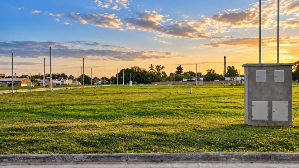 Altos de San Isidro al atardecer: lotes con pasto, calle con cordón, postes de alumbrado y el gabinete de medidores, con casas de Lules al fondo